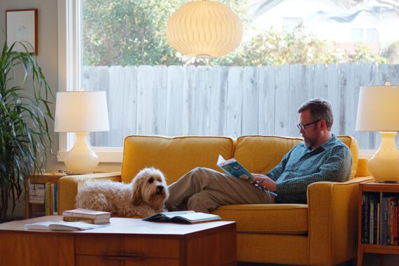 A man reading a book on a couch next to a dog